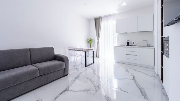 A modern living room with a grey sofa, a dining table with chairs, a kitchen with white cabinets, and a large window with curtains. - Alberti Apartments Historical Center Trapani (Trapani)
