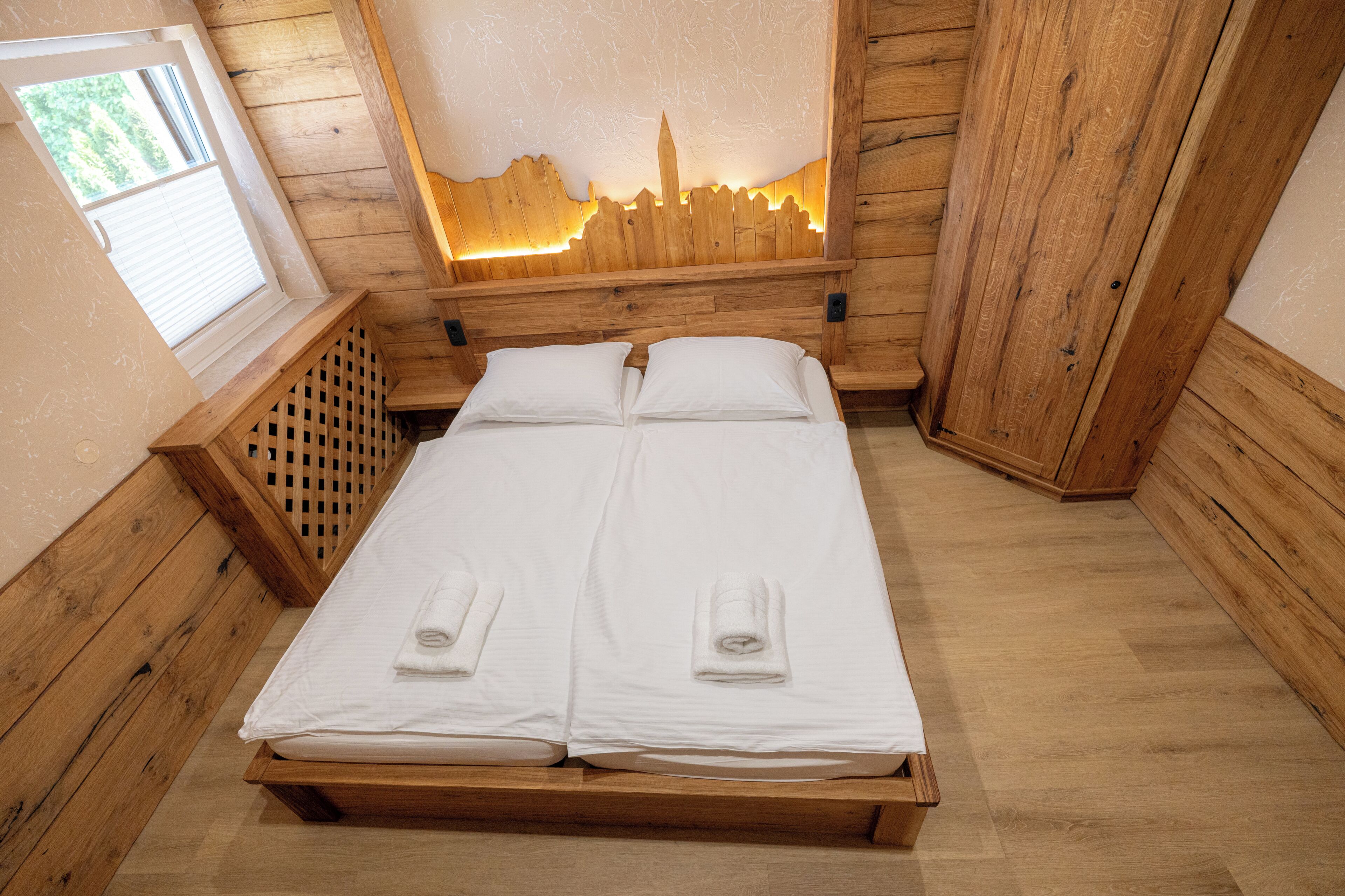 Un lit superposé en bois avec deux draps et deux serviettes blancs, une tête de lit en bois ornée d’un motif lumineux représentant un paysage urbain, et une fenêtre avec des stores.