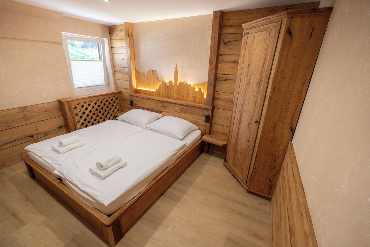 A wooden-framed bed with white linens and towels, a window with blinds, and a wall-mounted wooden cabinet.
