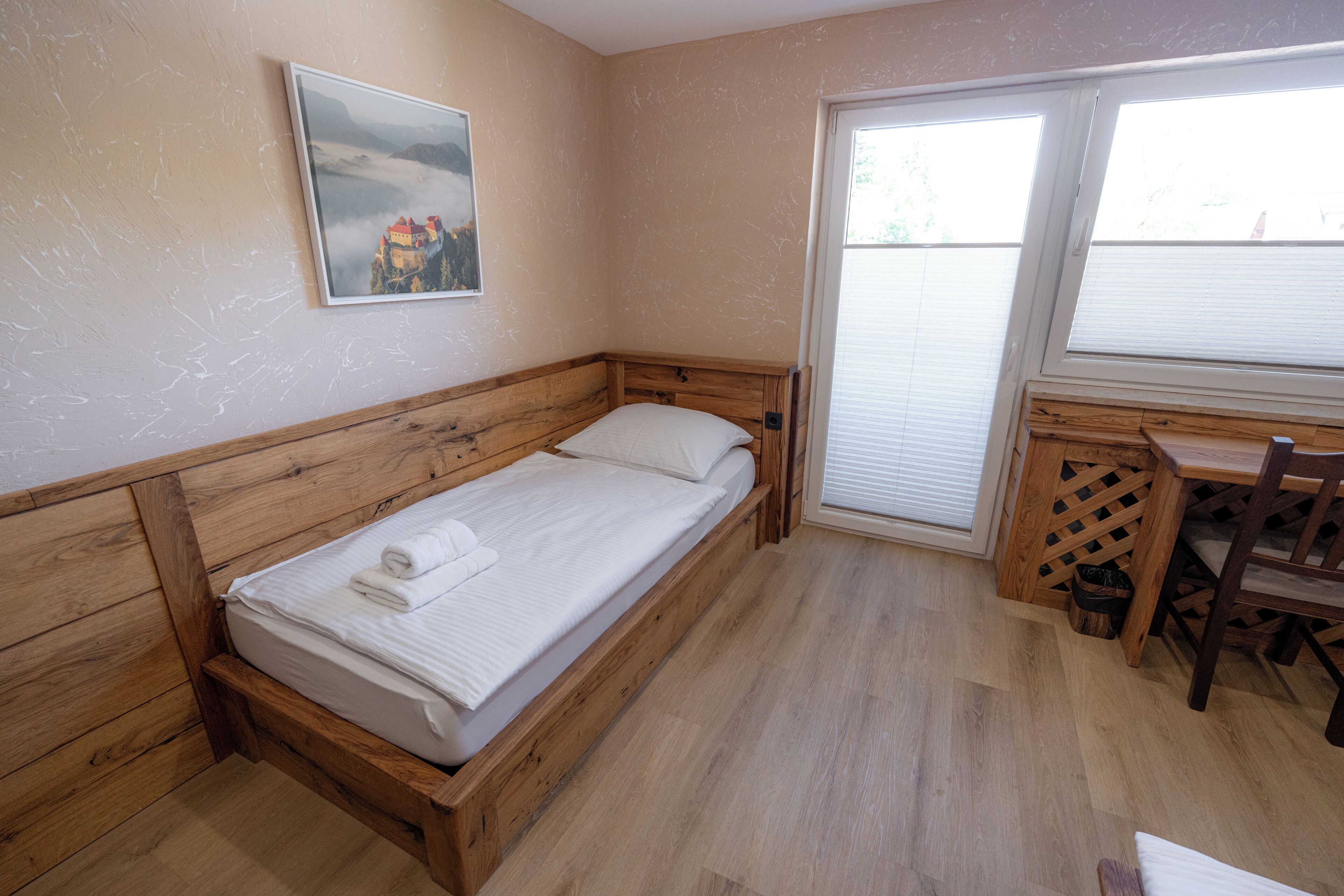 A single bed with a wooden headboard, a small desk with a chair, and a window with blinds.