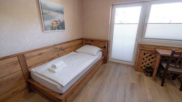 A single bed with a wooden headboard, a small desk with a chair, and a window with blinds.