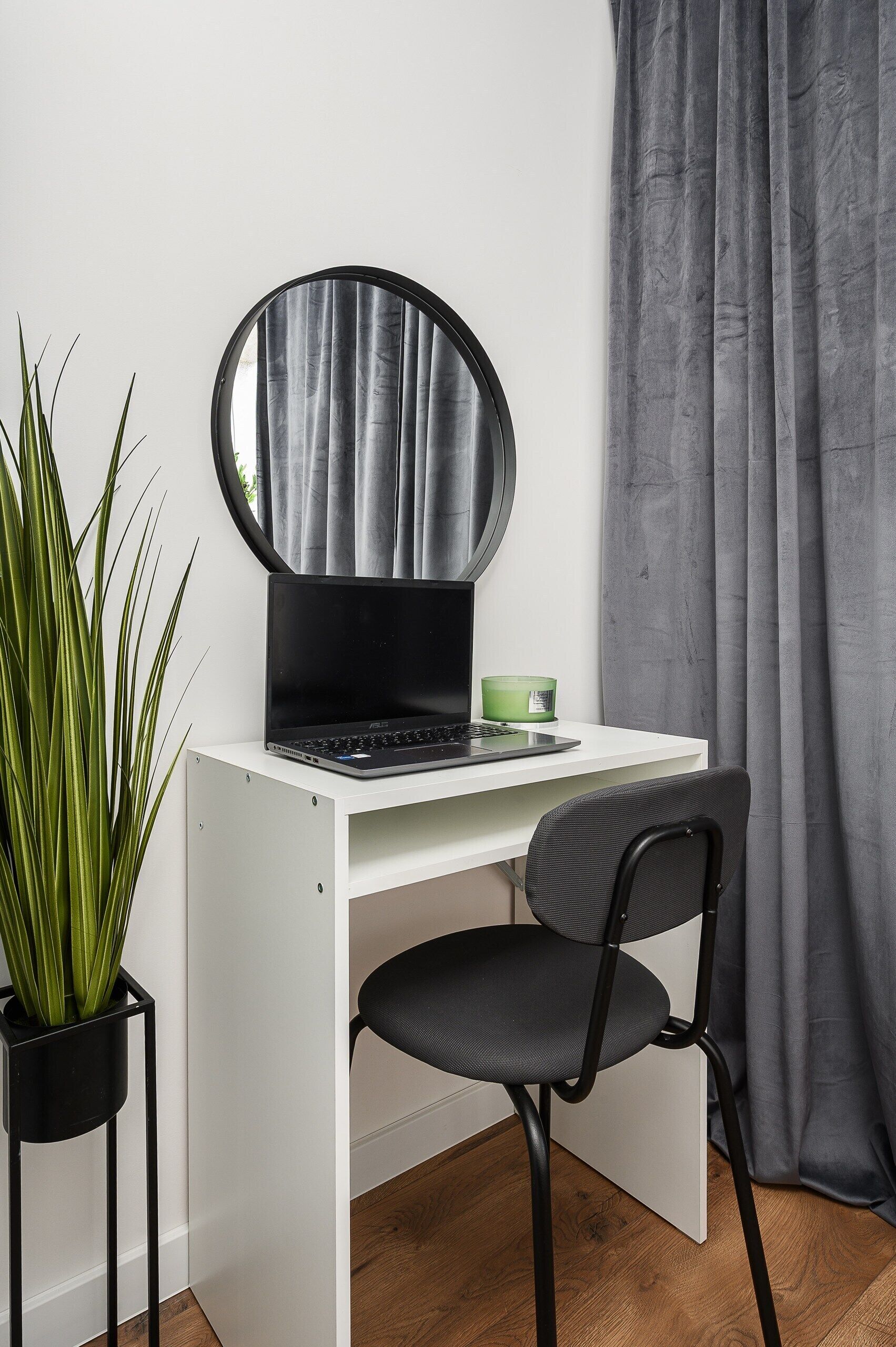 Un bureau blanc avec un ordinateur portable, une tasse verte, un miroir rond, une chaise noire et une plante en pot.