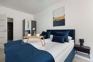 Een moderne slaapkamer met een groot bed, een nachtkastje met een lamp, een klein tafeltje met kop en schotel, en een schilderij aan de muur.