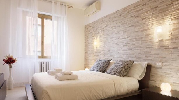 A modern bedroom with a large bed, white linens, a brick-patterned wall, and a bedside table with a lamp. - Bilocale moderno a Bollate vicino a Fieramilano e Milano Ice Park (Bollate)