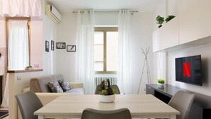 A modern living room with a dining table, grey chairs, a grey sofa, a TV mounted on the wall, and a window with white curtains. - Bilocale moderno a Bollate vicino a Fieramilano e Milano Ice Park (Bollate)