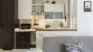 A compact kitchen with a microwave, sink, and cabinets, a gray sofa with a patterned pillow, and a window with a view. - Bilocale moderno a Bollate vicino a Fieramilano e Milano Ice Park (Bollate)