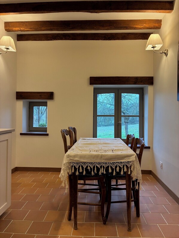 A dining room with a table set for four, wooden chairs, two wall-mounted lamps, and two windows with views of trees. - Les Peupliers du Perche, a charming farmstay in Normandy surrounded by nature (Parfondeval)