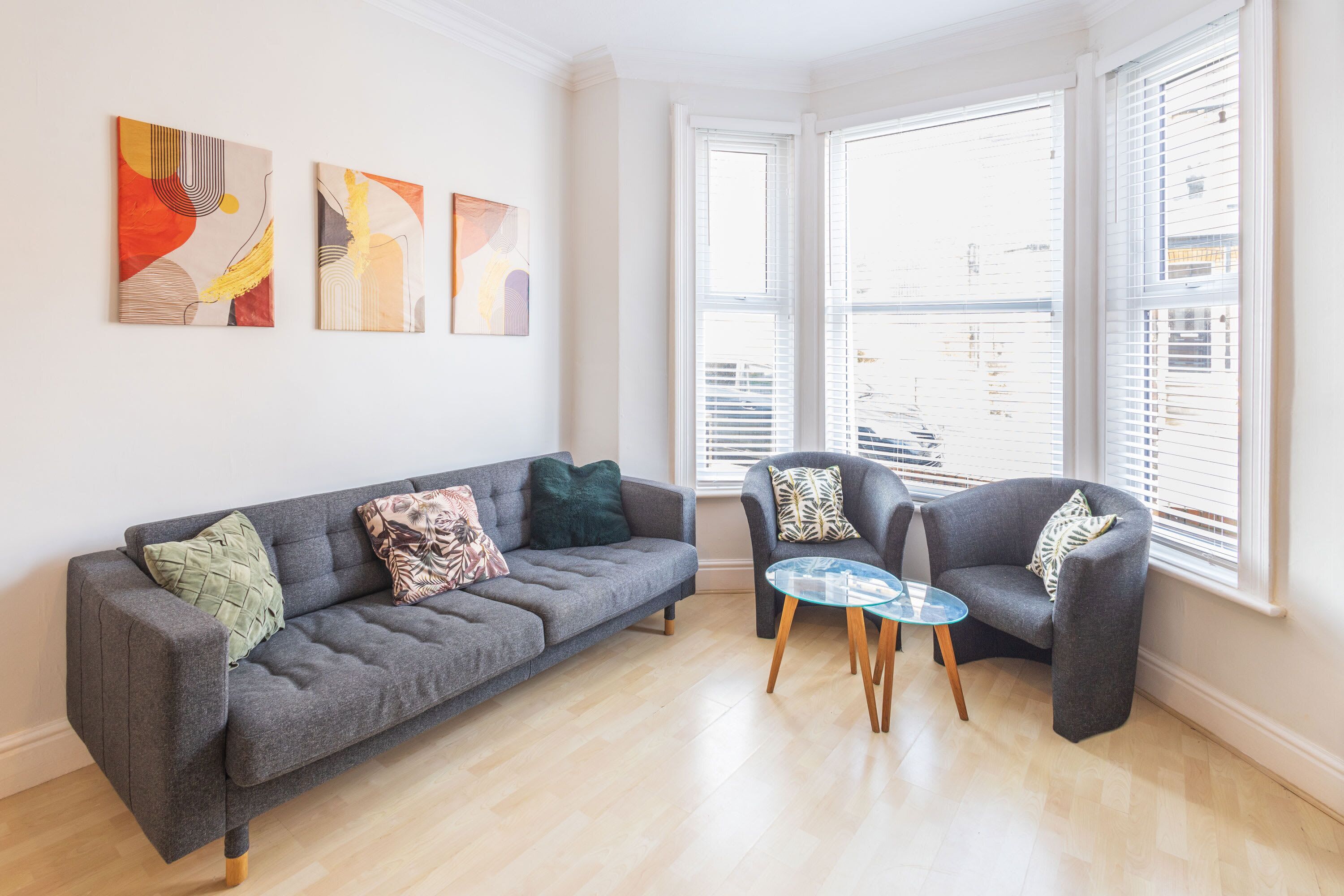 A modern living room with a grey sofa, two armchairs, a glass coffee table, and abstract wall art.