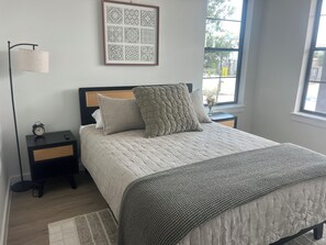 Une chambre à coucher avec un lit, une table de chevet avec un réveil, une lampe de chevet, une fenêtre donnant sur un paysage agréable et une œuvre d’art encadrée accrochée au mur.
