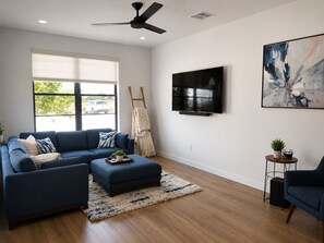 A modern living room with a sectional sofa, a small ottoman, a flat-screen TV, and a painting on the wall. - Brand new 2-bedroom condo in beautiful New Braunfels (New Braunfels)