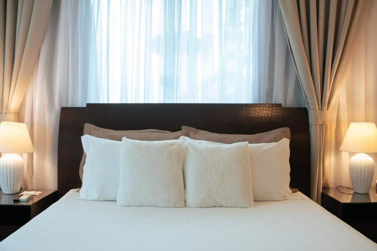 A neatly made bed with white linens and two pillows, flanked by two bedside tables with lamps, in a room with sheer curtains and a dark headboard.