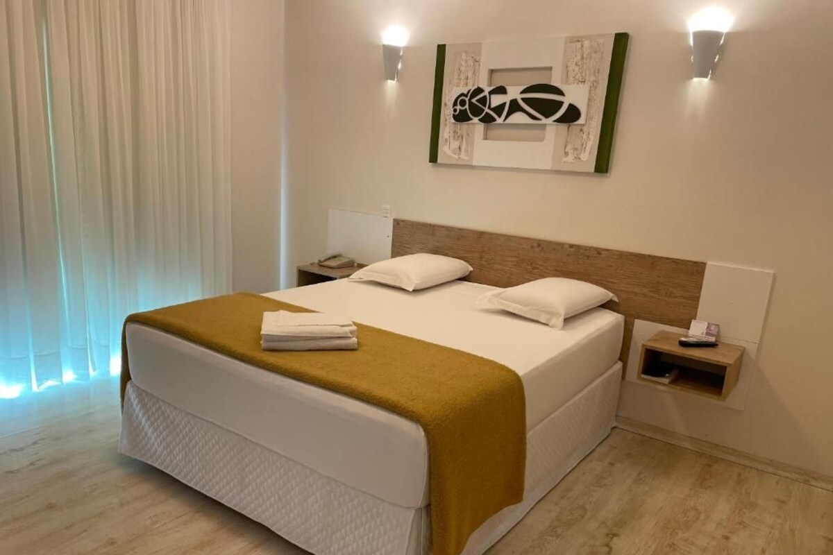 A hotel room with a bed, bedside tables, a wall-mounted artwork, and a window with curtains.