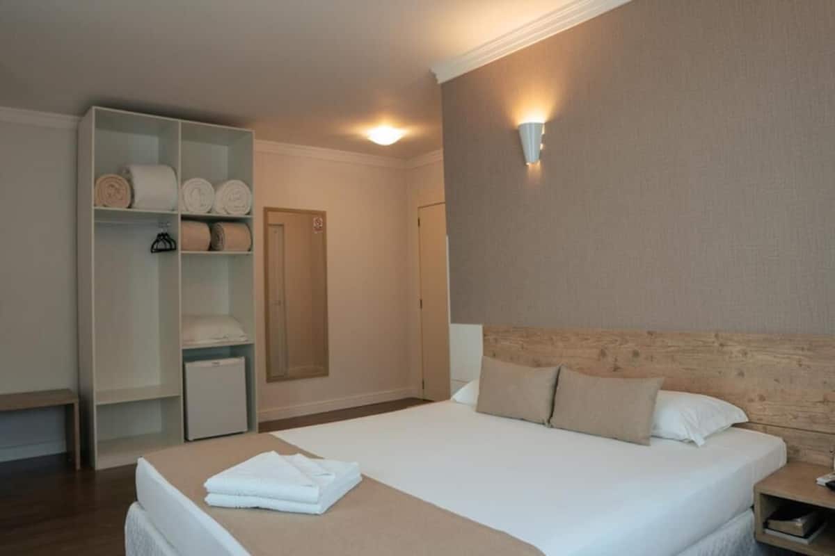 A hotel room with a bed, a wardrobe, a bedside table, and a wall-mounted light fixture.