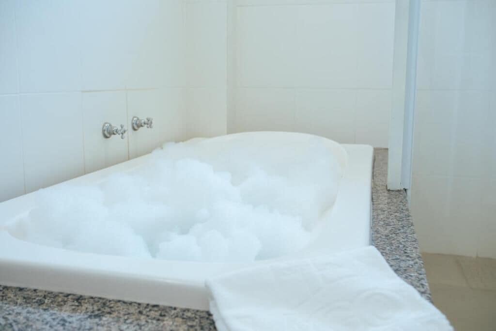 A bathtub filled with white bath products, a granite countertop, and white tiled walls.