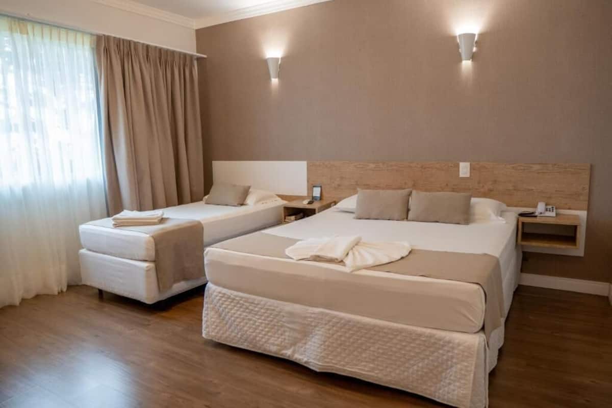 A hotel room with two beds, a wooden headboard, a bedside table with a lamp, and a window with curtains.