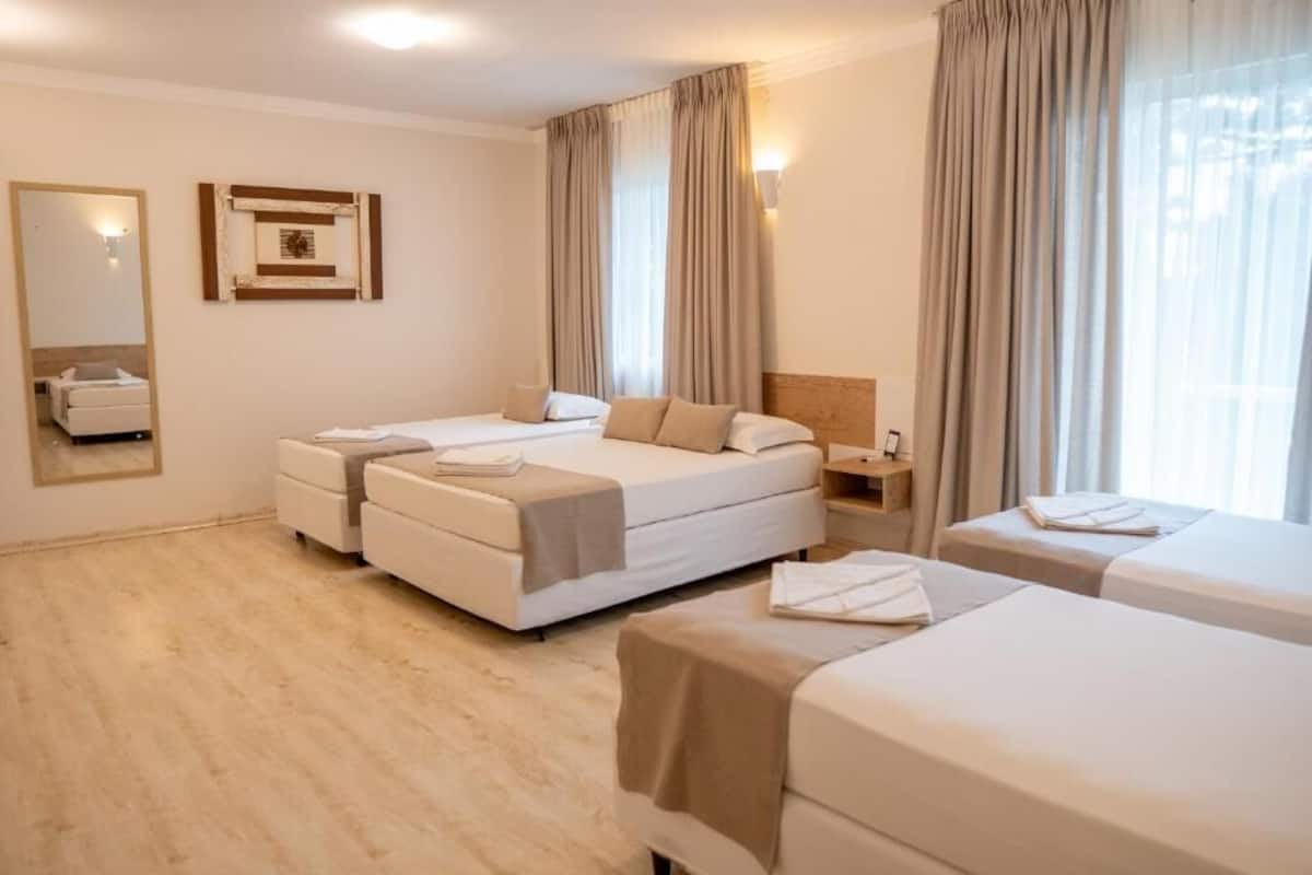 A hotel room with a large bed, two smaller beds, a bedside table, a mirror, and a window with curtains.