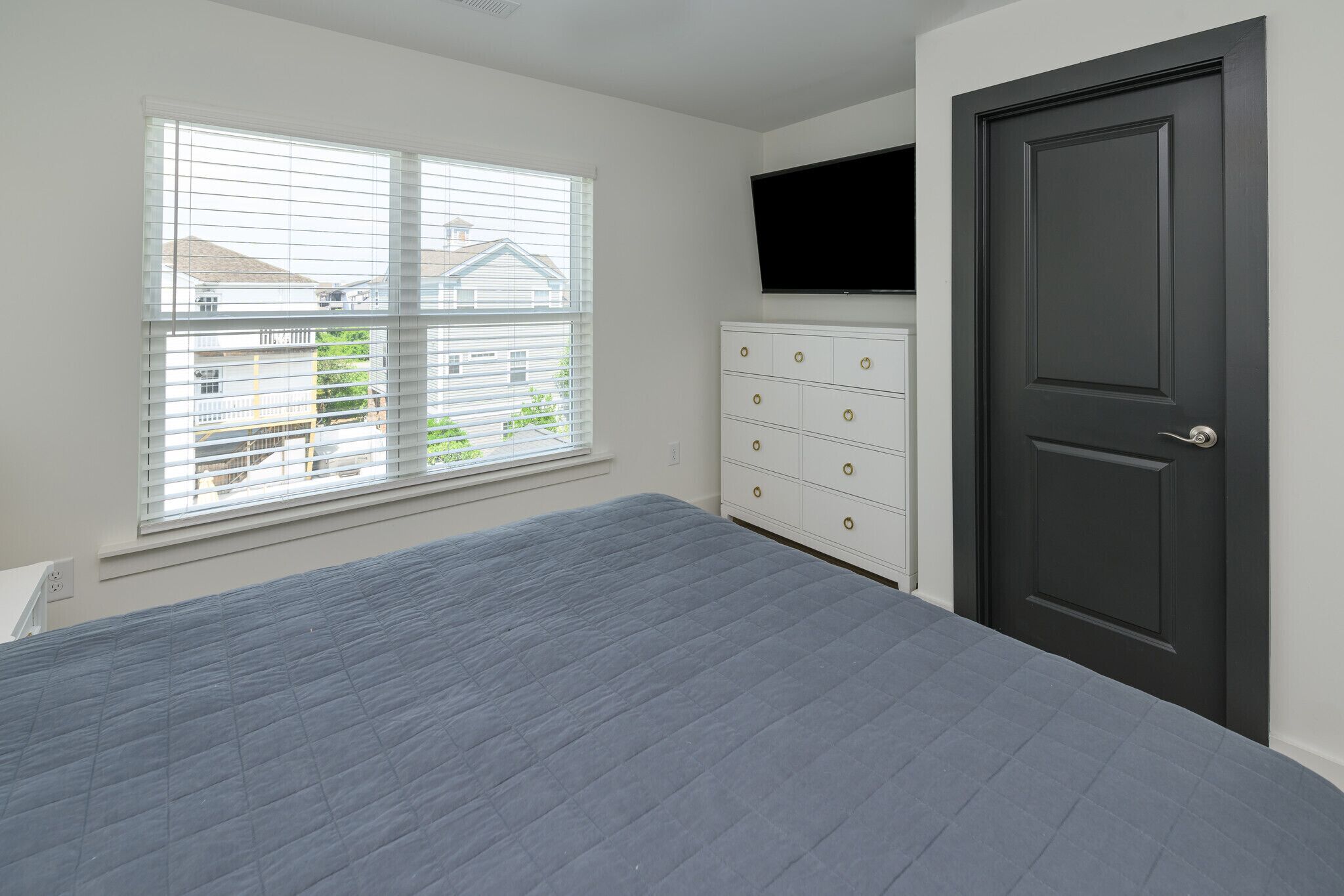 A bedroom with a bed, a dresser, a TV mounted above a door, and a window with blinds.