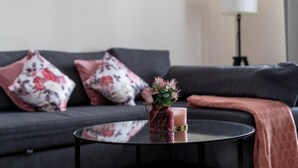 Un salon moderne avec un canapé gris, des coussins à motifs floraux, une table basse en verre et un vase rempli de fleurs.