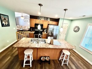 A kitchen with wooden cabinets, a granite countertop, stainless steel appliances, and two white bar stools.