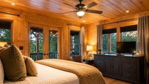 Ein Schlafzimmer in einer Holzhütte mit einem Bett, einem Deckenventilator, einem Fernseher auf einer Kommode und großen Fenstern.