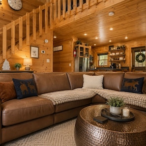 Ein gemütliches Blockhaus-Interieur mit einer braunen Ledersofa, einem runden Couchtisch, Holzwänden, einer Küchenzeile und einer Treppe.