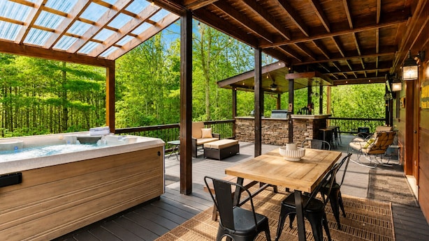Ein überdachter Außenbereich mit Whirlpool, Holzterrasse, Sitzmöbeln und Kamin aus Stein.