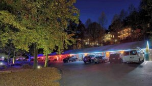 Un espace de stationnement avec plusieurs véhicules, un bâtiment dont les fenêtres sont illuminées et un arbre au premier plan.