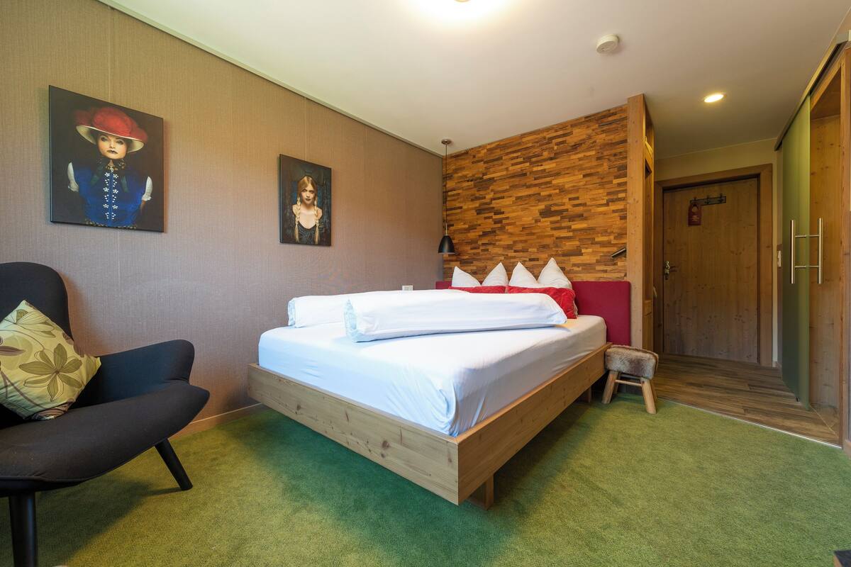 A bedroom with a bed, a chair, a small stool, and two paintings on the wall.