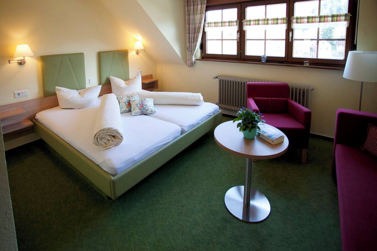 A hotel room with a bed, a small round table, a red armchair, and a window with curtains.