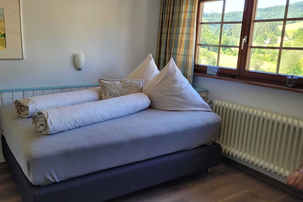 A bed with rolled towels, a radiator, a window with a view, and a painting on the wall.