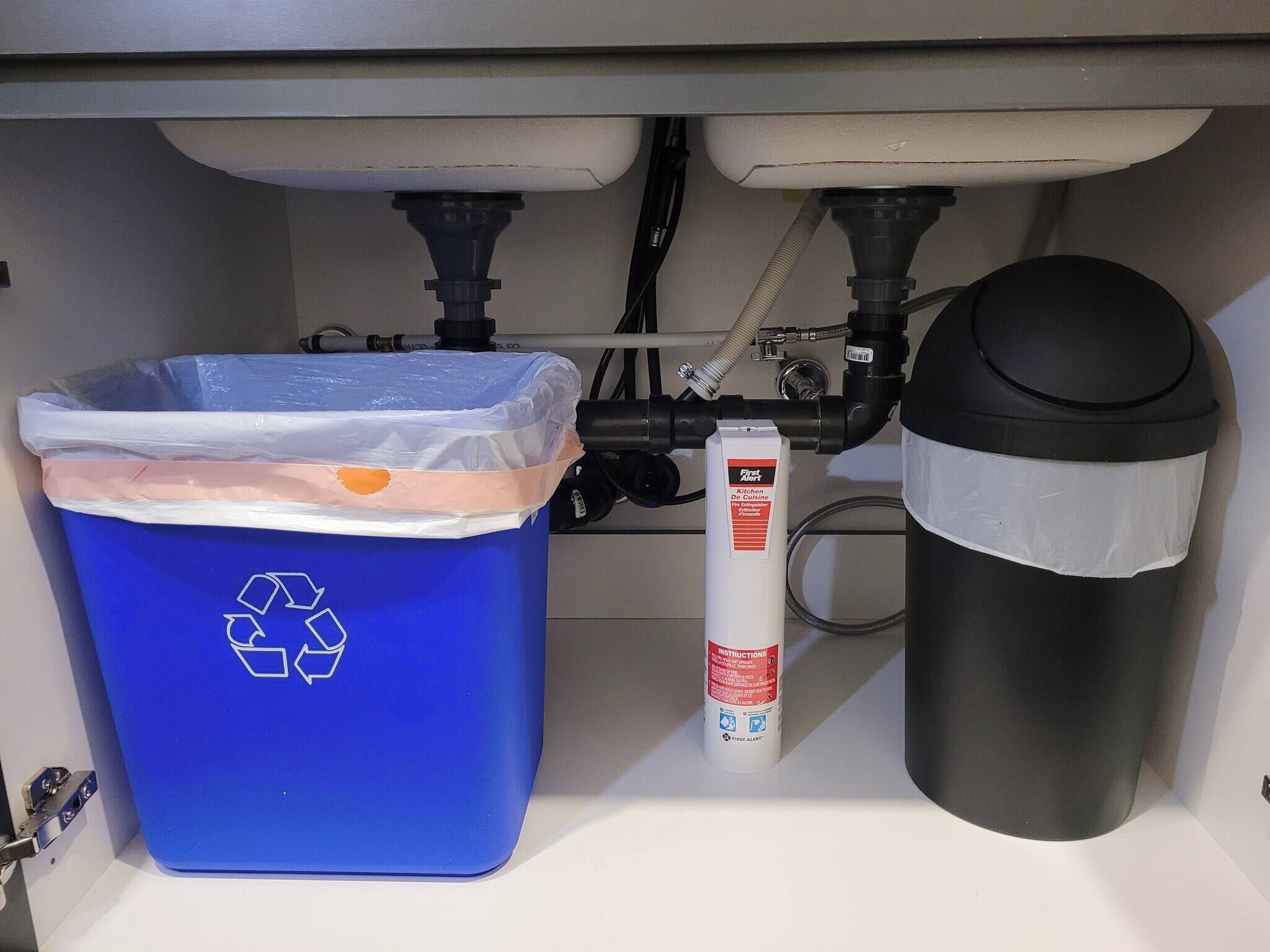 Ein Küchenbereich mit einer Spüle, einer blauen Recyclingtonne, einer schwarzen Mülltonne und einem weißen First Alert Feuerlöscher.