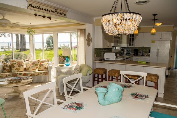 A spacious living area with a large window overlooking a garden, a dining table with chairs, and a kitchen area with white cabinets and a chandelier. - Lovely 3 BR, Rappahannock Riverfront Retreat in Picturesque Urbanna (Urbanna)