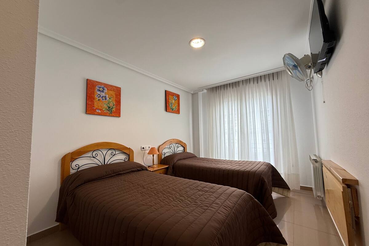 A hotel room with two beds, a ceiling fan, a TV mounted on the wall, and two paintings on the wall.