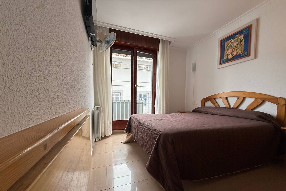 A bedroom with a wooden headboard, a bed with a brown bedspread, a radiator, a window with curtains, and a painting on the wall.