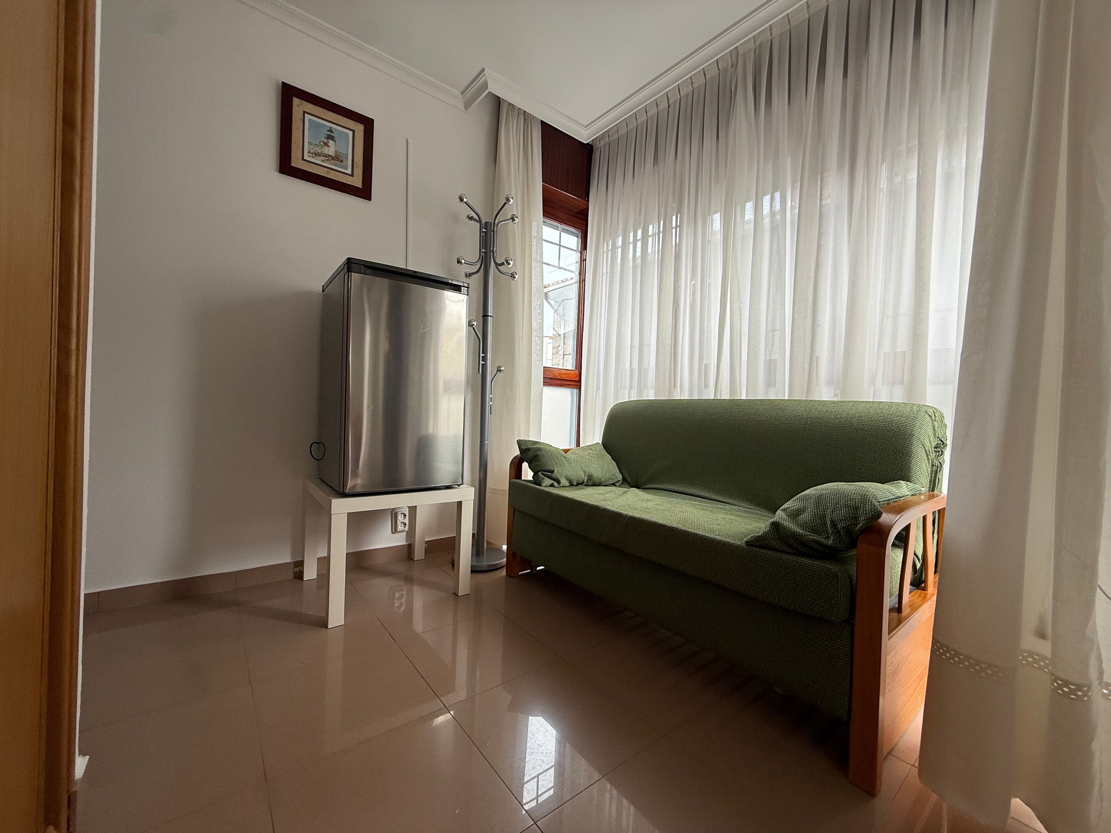Una sala de estar con un sofá verde, un refrigerador independiente, una silla de madera, un cuadro en la pared y una ventana con cortinas.