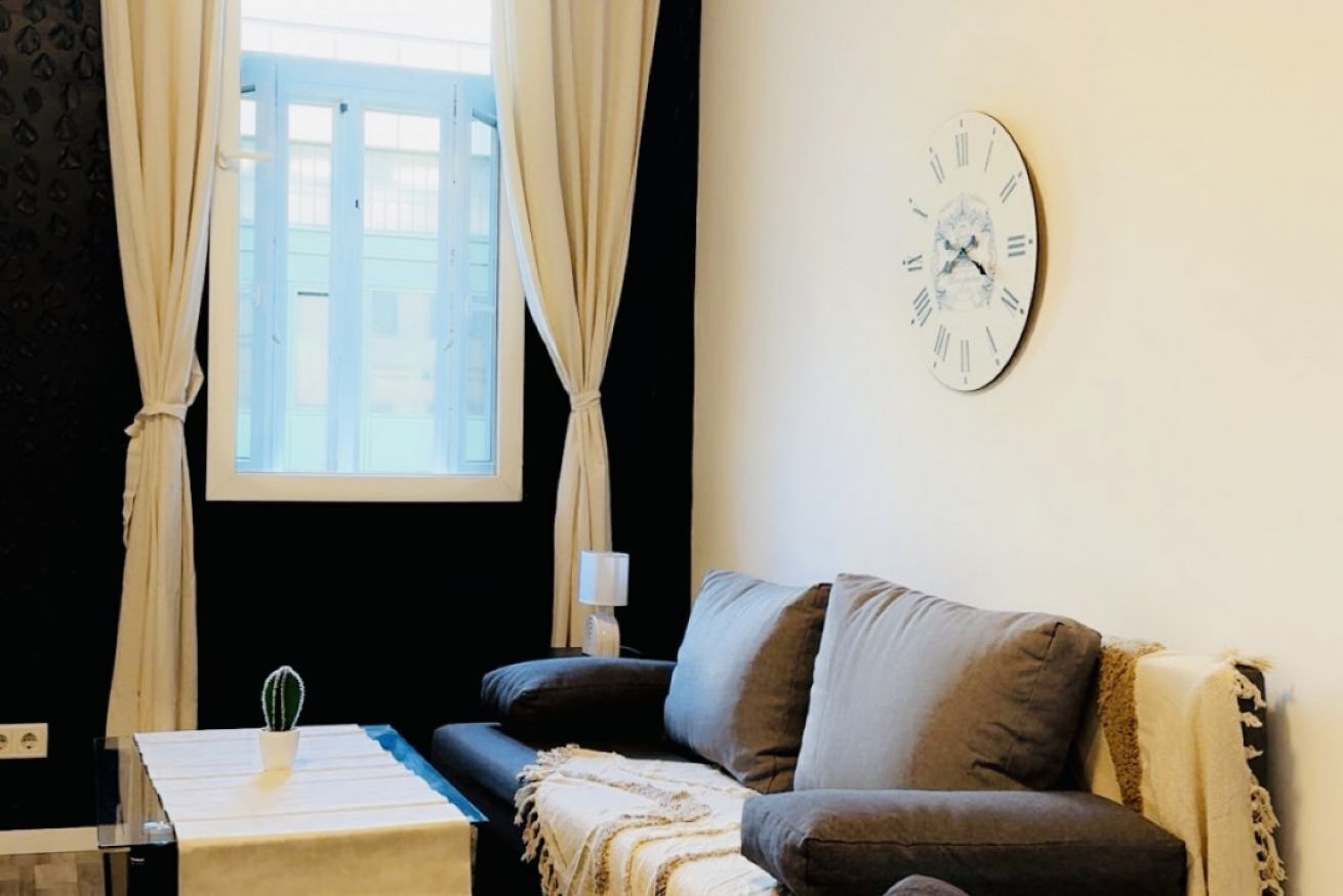 Une pièce avec un canapé, une petite table sur laquelle se trouve un cactus, une horloge au mur et une fenêtre avec des rideaux.