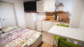 Un dormitorio compacto con una colcha colorida, una kitchenette pequeña con microondas y un televisor sobre los armarios.