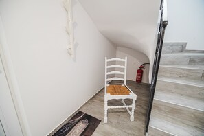 Valkoinen tuoli, jonka istuin on punottu, seisoo käytävällä, jossa on puulattia, palosammutin ja puuportaat.