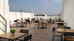 An outdoor dining area with multiple tables and chairs, a staircase on the left, and a view of the cityscape in the background. - Hotel Golden Palace (Agra)