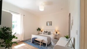 Une chambre à coucher avec un lit, des tables de chevet, un bureau et une chaise. Il y a une fenêtre avec des rideaux, une plante et un luminaire au plafond.