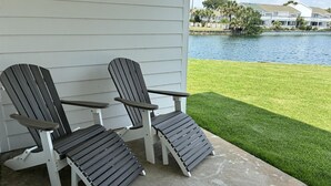 Zwei Adirondack-Stühle auf einer Beton-Terrasse mit Blick auf einen See und Häuser im Hintergrund.
