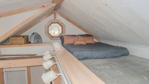 Ein kleines Schlafzimmer im Dachgeschoss mit schrägen Wänden, einem Bett mit Kissen, einer Holzbank und einer Pendelleuchte.