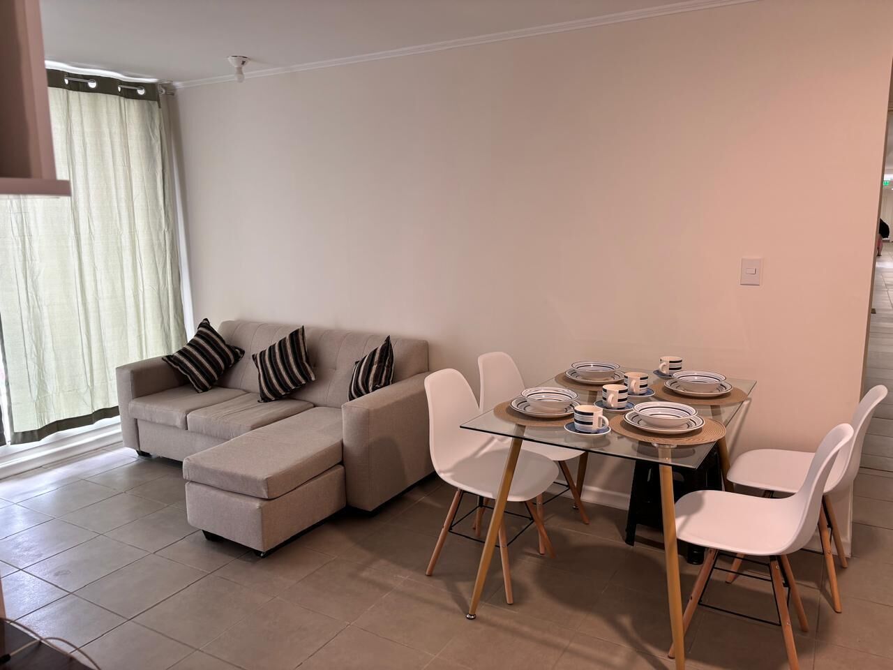 A modern living room with a beige sectional sofa, a glass dining table with white chairs, and a large window with curtains.