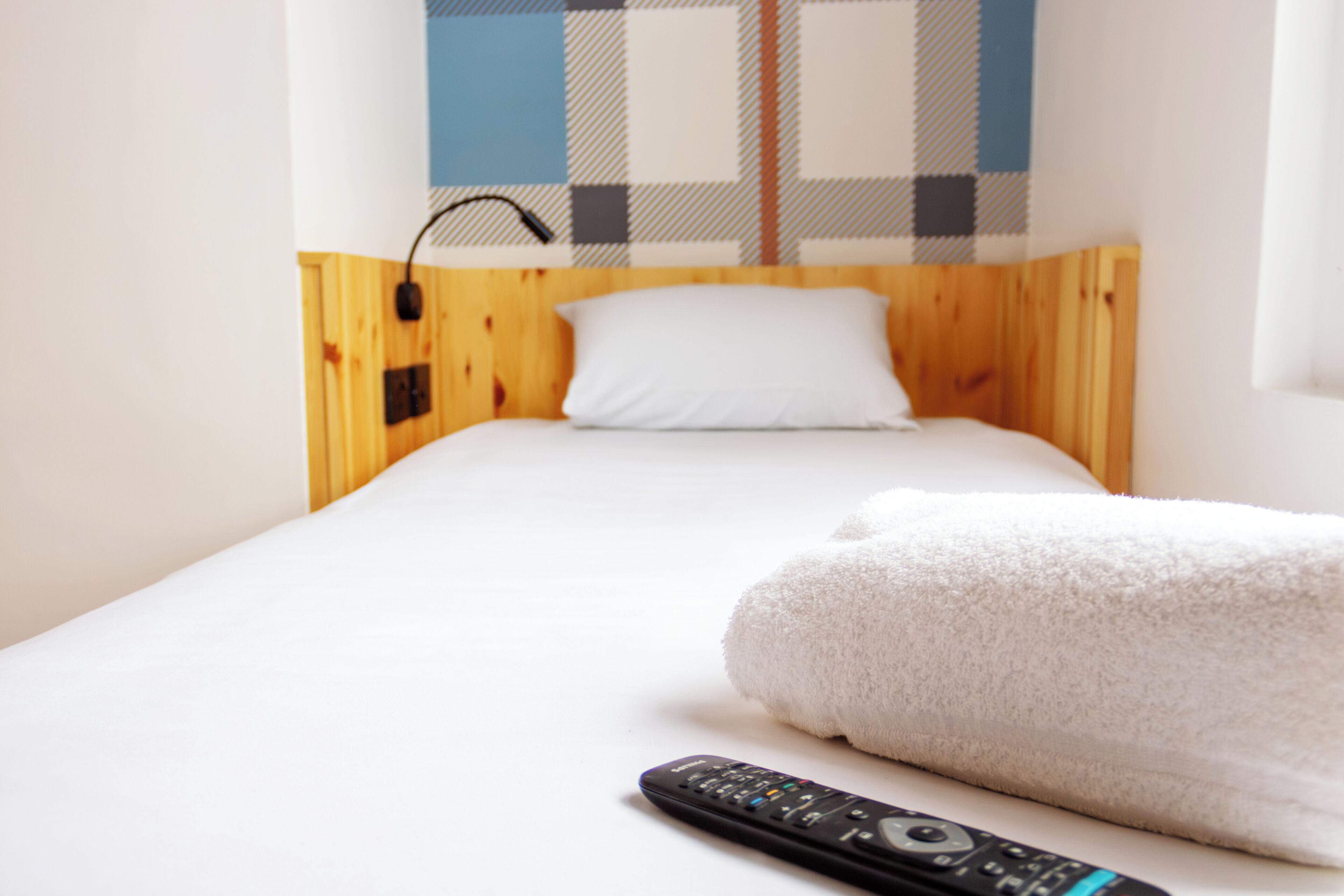 A single bed with a white pillow and a blanket, a remote control on the bed, and a checkered wall behind.