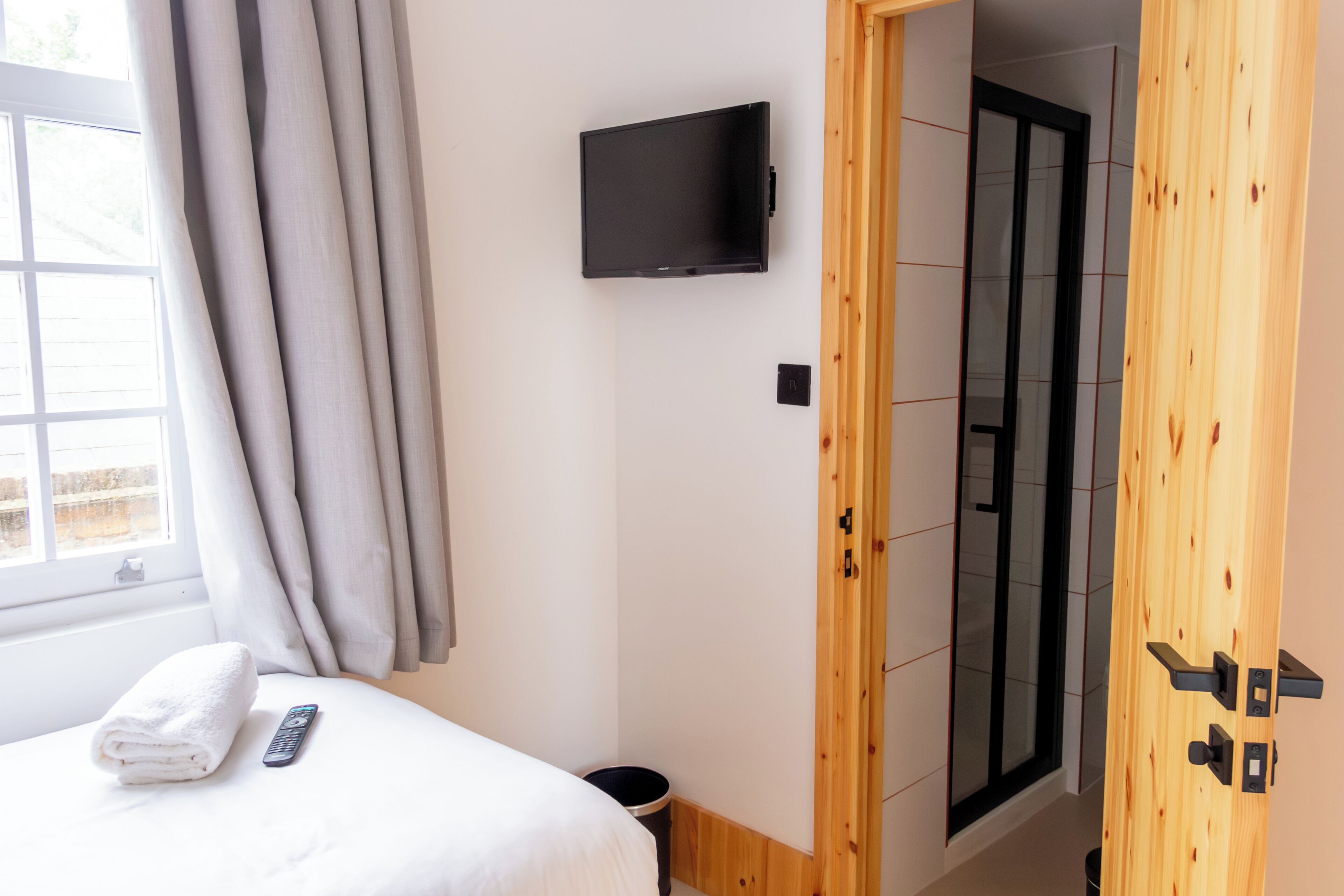 Ein Hotelzimmer mit einem Bett, einer Fernbedienung, einem an der Wand befestigten Fernseher, einem Fenster mit Vorhängen und einer Holztür.