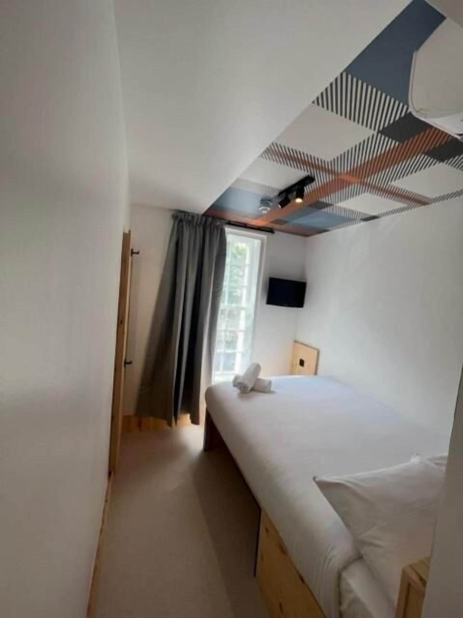 A bedroom with a bed, a window with curtains, a flat-screen TV on the wall, and a ceiling with a geometric pattern.