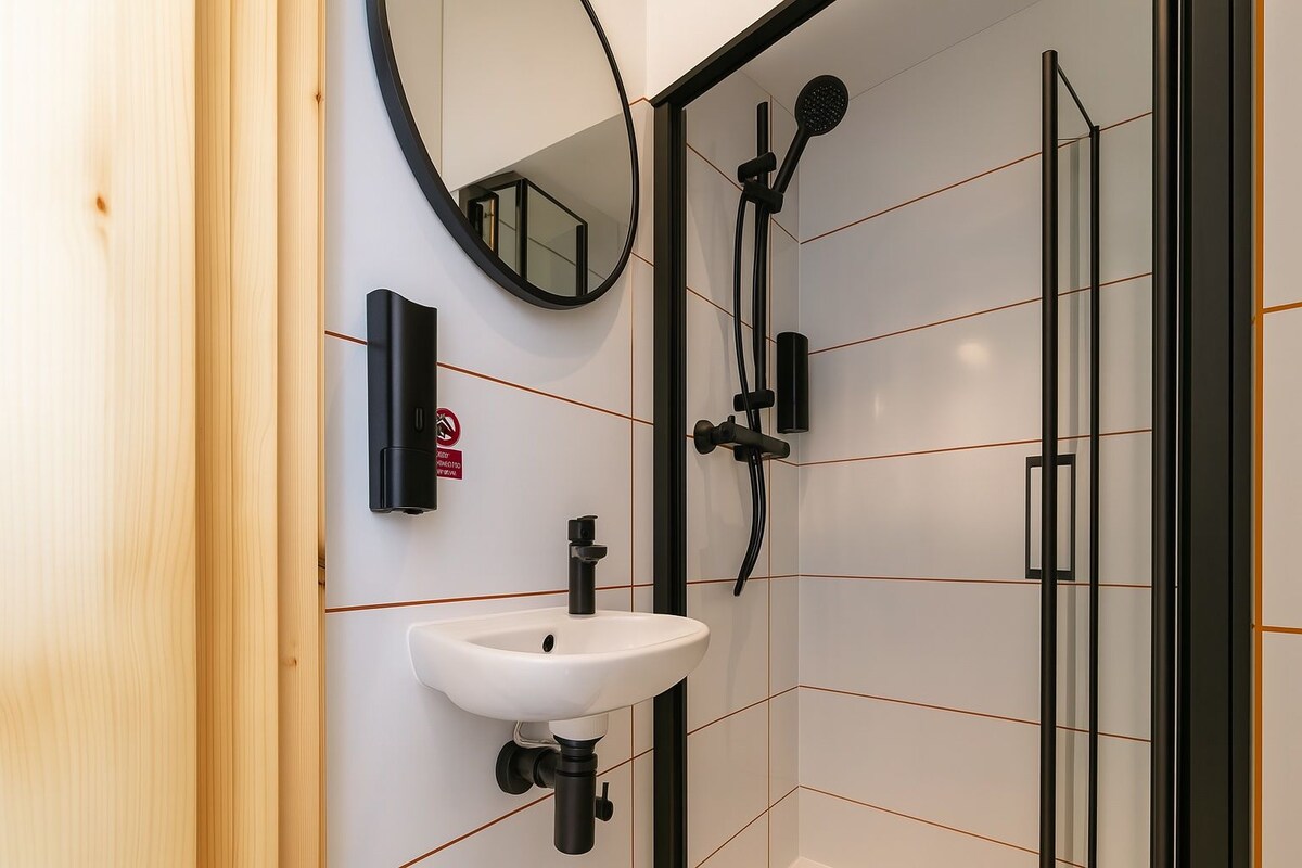 Un baño con lavamanos blanco, un espejo, ducha con cabezal de ducha manual y puerta de vidrio para la ducha.