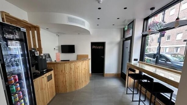 A modern hotel lobby with a wooden reception desk, a private room sign, a vending machine, and a seating area with black chairs. - EasyHotel Paddington (London)