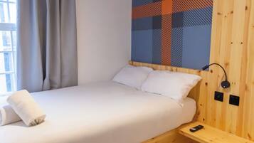 Una cama individual con ropa de cama blanca, cabecera de madera, mesita de noche pequeña con un control remoto y un enchufe en la pared.