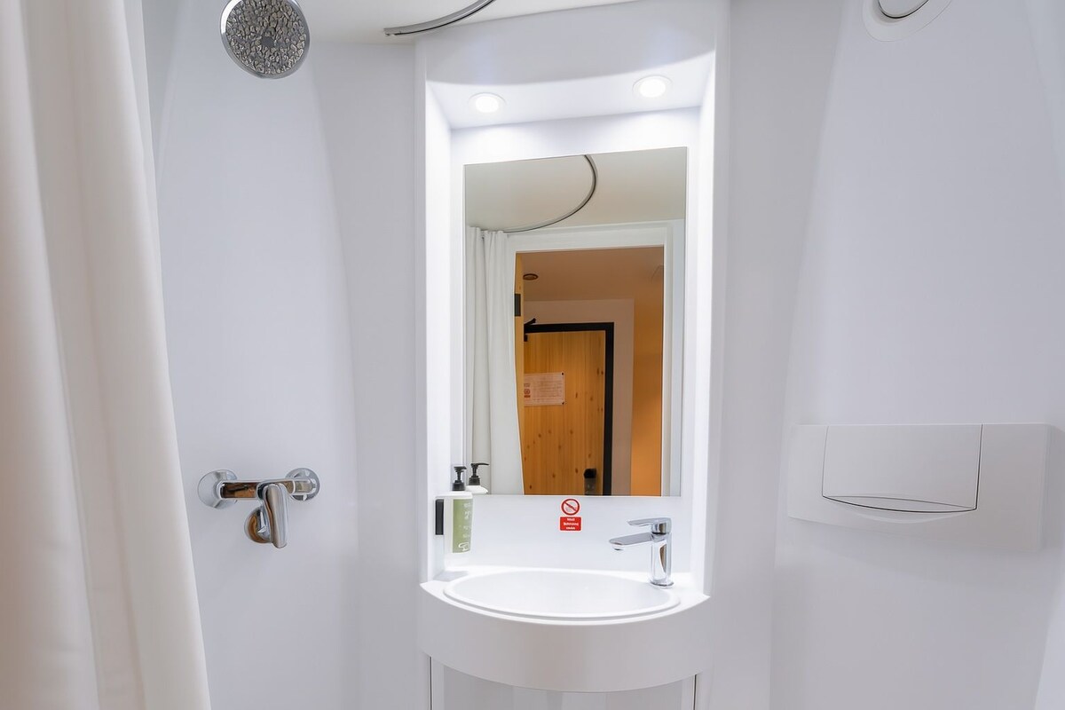 A bathroom with a white sink, a mirror, a showerhead, and a door with a sign.